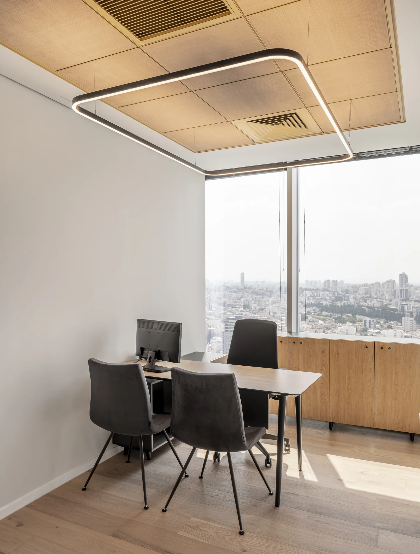 An office with a suspended rectangular ring luminaire over the desk and a city view